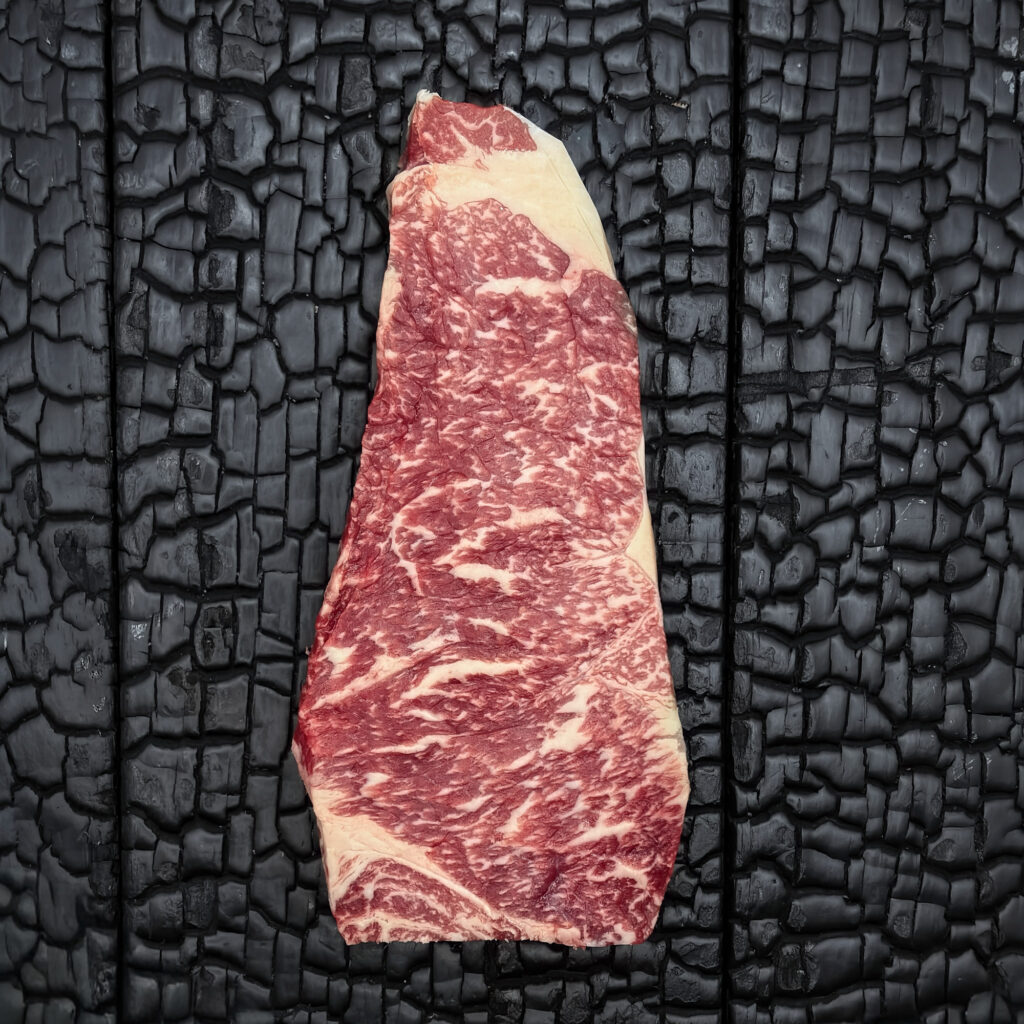 Rohes Striploin-Steak vom Wagyu-Kreuzungsrind mit ausgeprägter Marmorierung, rote Farbe mit gleichmäßigem Fettrand, mittig auf dunkler, verkohlter Holzoberfläche.