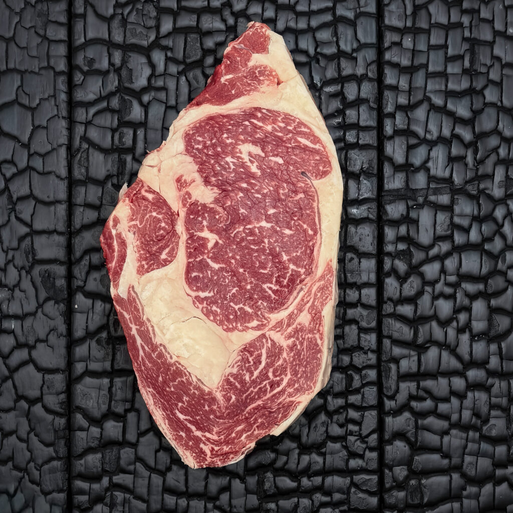 Rohes Rib-Eye-Steak vom Wagyu-Kreuzungsrind mit guter Marmorierung, rote Farbe mit deutlich sichtbarem Fettauge und Fettrand, mittig auf dunkler, verkohlter Holzoberfläche.