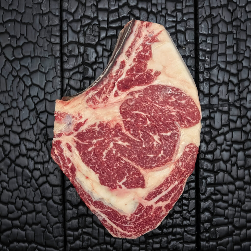 Rohes Rib-Eye-Steak mit Knochen vom Wagyu-Kreuzungsrind, gute Marmorierung, rote Farbe mit sichtbarem Fettauge und Fettrand, mittig auf dunkler, verkohlter Holzoberfläche.