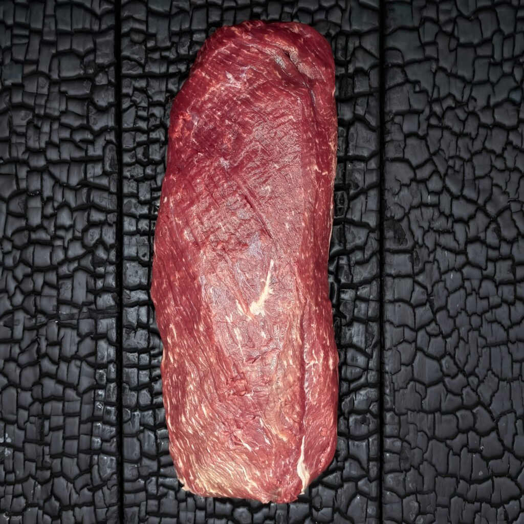 Ganzes rohes Rumpfilet vom Wagyu-Kreuzungsrind, längliche Form, feine Marmorierung, rote Farbe, mittig auf dunkler, verkohlter Holzoberfläche.