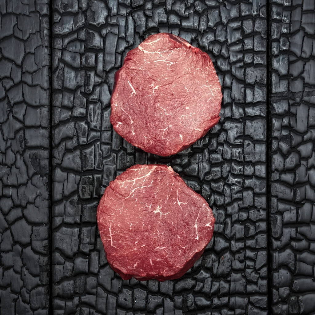 Zwei rohe Rumpfilet-Medaillons vom Wagyu-Kreuzungsrind, fein marmoriert, rote Farbe, übereinander angeordnet auf dunkler, verkohlter Holzoberfläche.