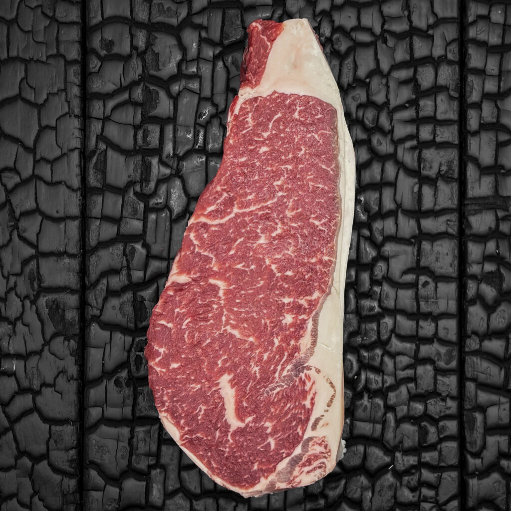 Rohes Striploin-Steak vom Wagyu-Kreuzungsrind mit guter Marmorierung, rote Farbe mit gleichmäßigem Fettrand, mittig auf dunkler, verkohlter Holzoberfläche.