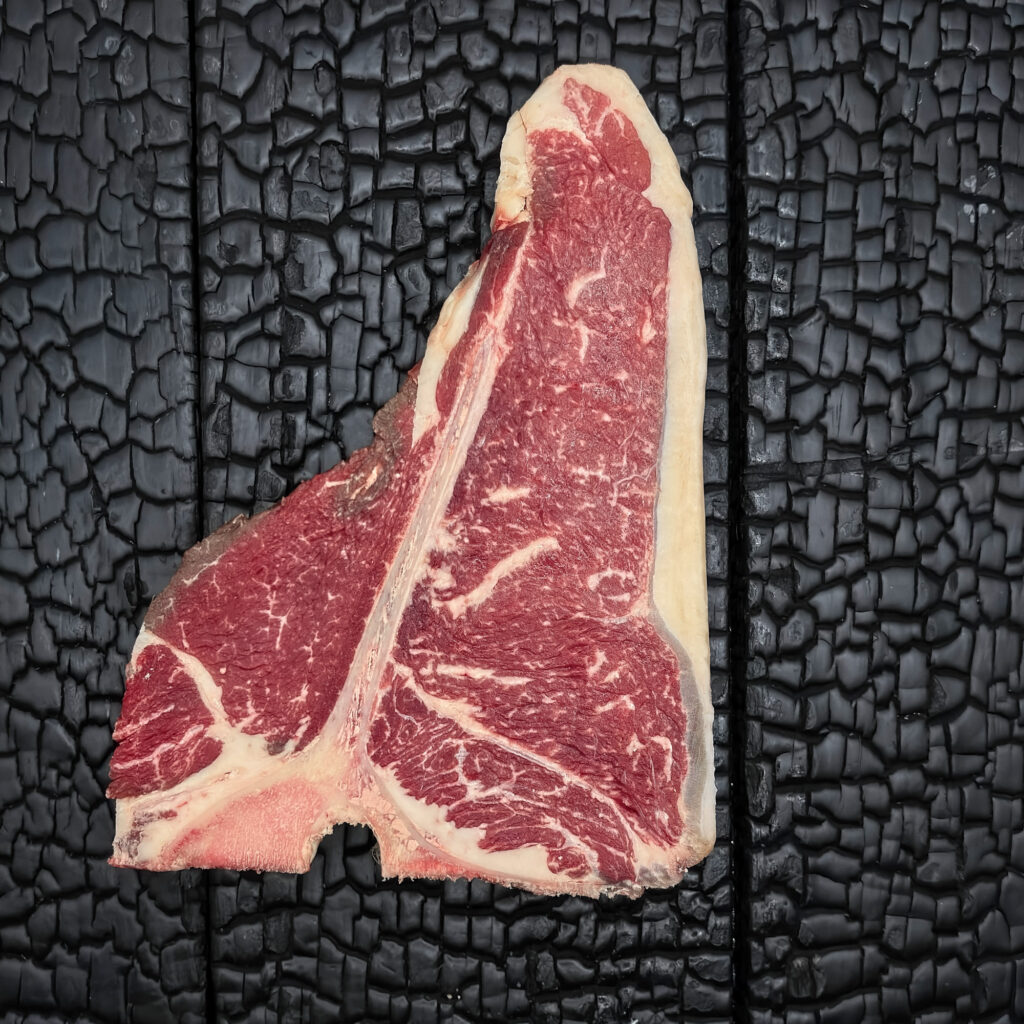 Rohes T-Bone-Steak mit T-förmigem Knochen vom Wagyu-Kreuzungsrind, gut marmoriert, Filet- und Beiriedanteil, rote Farbe mit hellem Fettrand, mittig auf dunkler, verkohlter Holzoberfläche.