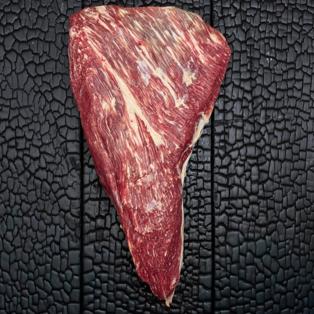 Rohes Tri-Tip im Ganzen vom Wagyu-Kreuzungsrind, dreieckige Form, gut marmoriert, rote Farbe mit Fettdeckel, mittig auf dunkler, verkohlter Holzoberfläche.