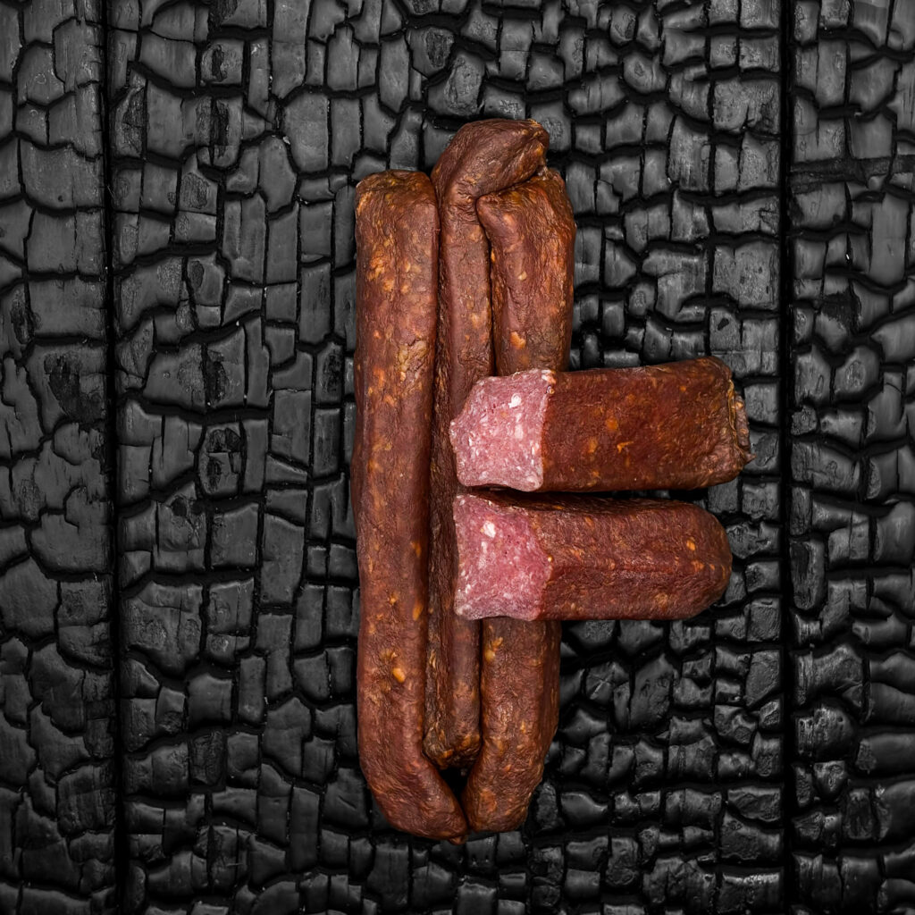 Mehrere Wagyu-Landjäger mit dunkelbrauner, leicht glänzender Wursthülle, parallel nebeneinander angeordnet. Zwei Stück sind quer angeschnitten, sodass das fein gekörnte, rötliche Innere sichtbar wird. Die Würstel liegen mittig auf einer dunklen, verkohlten Holzoberfläche.