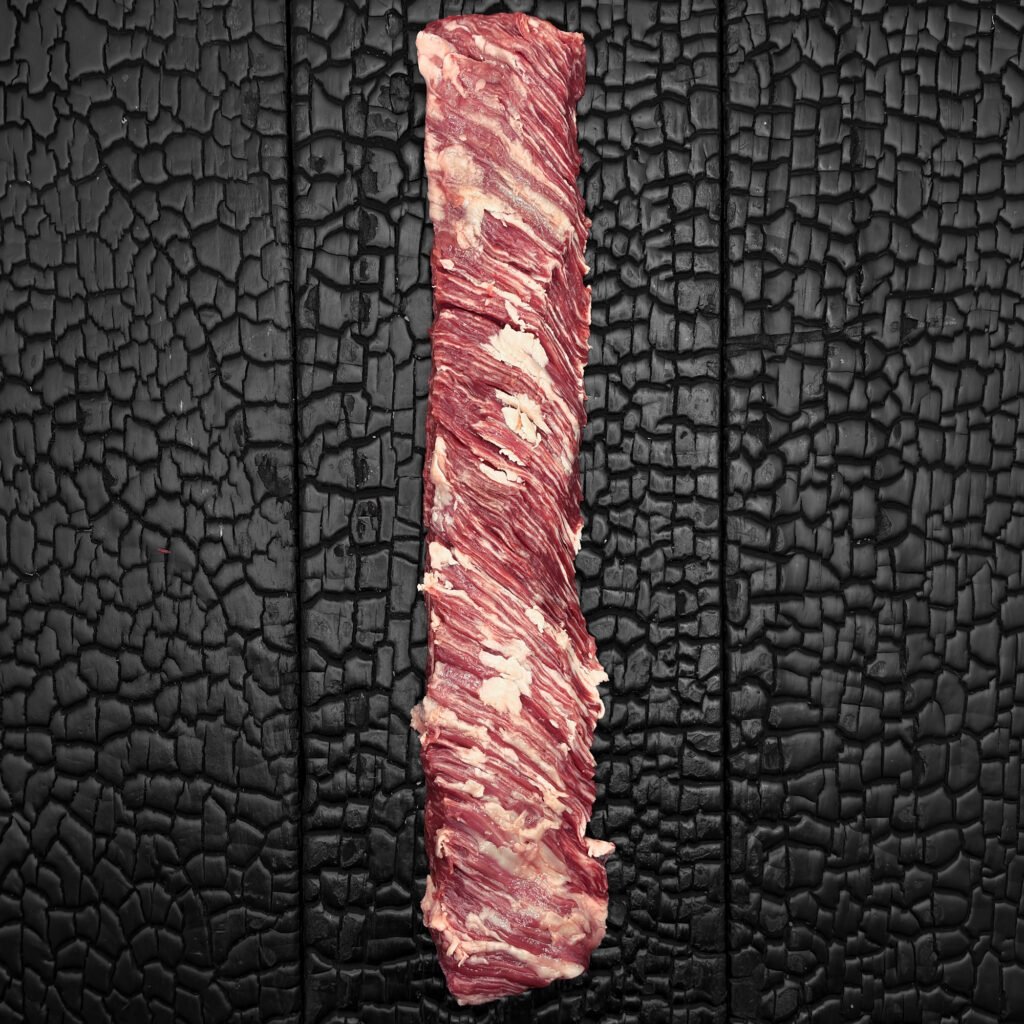 Rohes Outside Skirt vom Wagyu-Rind, stark marmoriert, rote Fleischfarbe mit grober Faserstruktur, langgezogenes Stück, mittig auf dunkler, verkohlter Holzoberfläche.