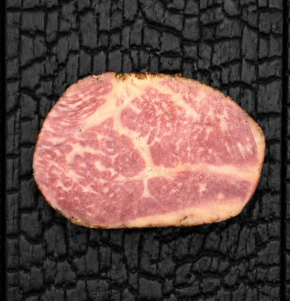 Ein einzelnes Stück Wagyu Pastrami liegt zentral auf einer schwarz verkohlten Holzoberfläche. Die ovale Scheibe zeigt eine feine helle Fettmarmorierung im rosafarbenen Fleisch sowie einen dunklen Gewürzrand. Die reduzierte Draufsicht und der starke Kontrast heben Struktur, Marmorierung und die hochwertige Fleischqualität hervor.