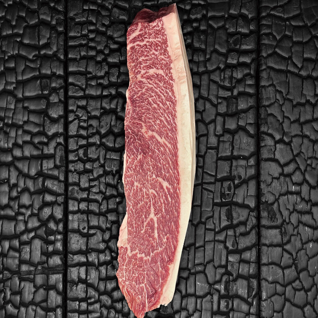 Rohes Picanha-Steak vom Wagyu-Rind, stark marmoriert, rote Fleischfarbe mit feiner Faserstruktur und deutlichem Fettrand, längliches Stück, mittig auf dunkler, verkohlter Holzoberfläche.