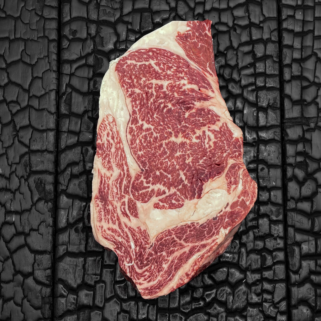 Rohes Rib-Eye-Steak vom Wagyu-Rind, stark marmoriert, rote Farbe mit ausgeprägtem Fettauge und Fettrand, mittig auf dunkler, verkohlter Holzoberfläche.