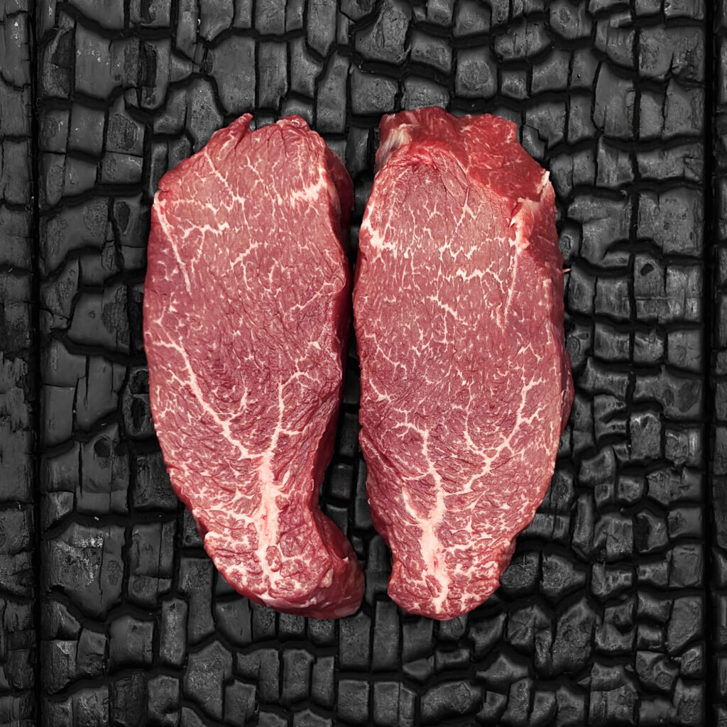 Zwei rohe Rumpfilet-Steaks vom Wagyu-Rind, gut marmoriert, rote Farbe, längliche Form, nebeneinander auf dunkler, verkohlter Holzoberfläche.