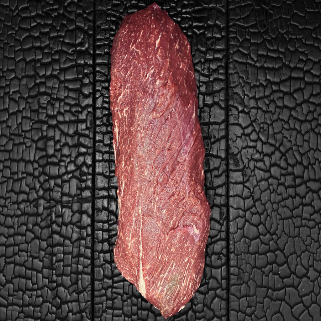 Ganzes rohes Rumpfilet vom Wagyu-Rind, längliche Form, gut marmoriert, rote Farbe, mittig auf dunkler, verkohlter Holzoberfläche.“