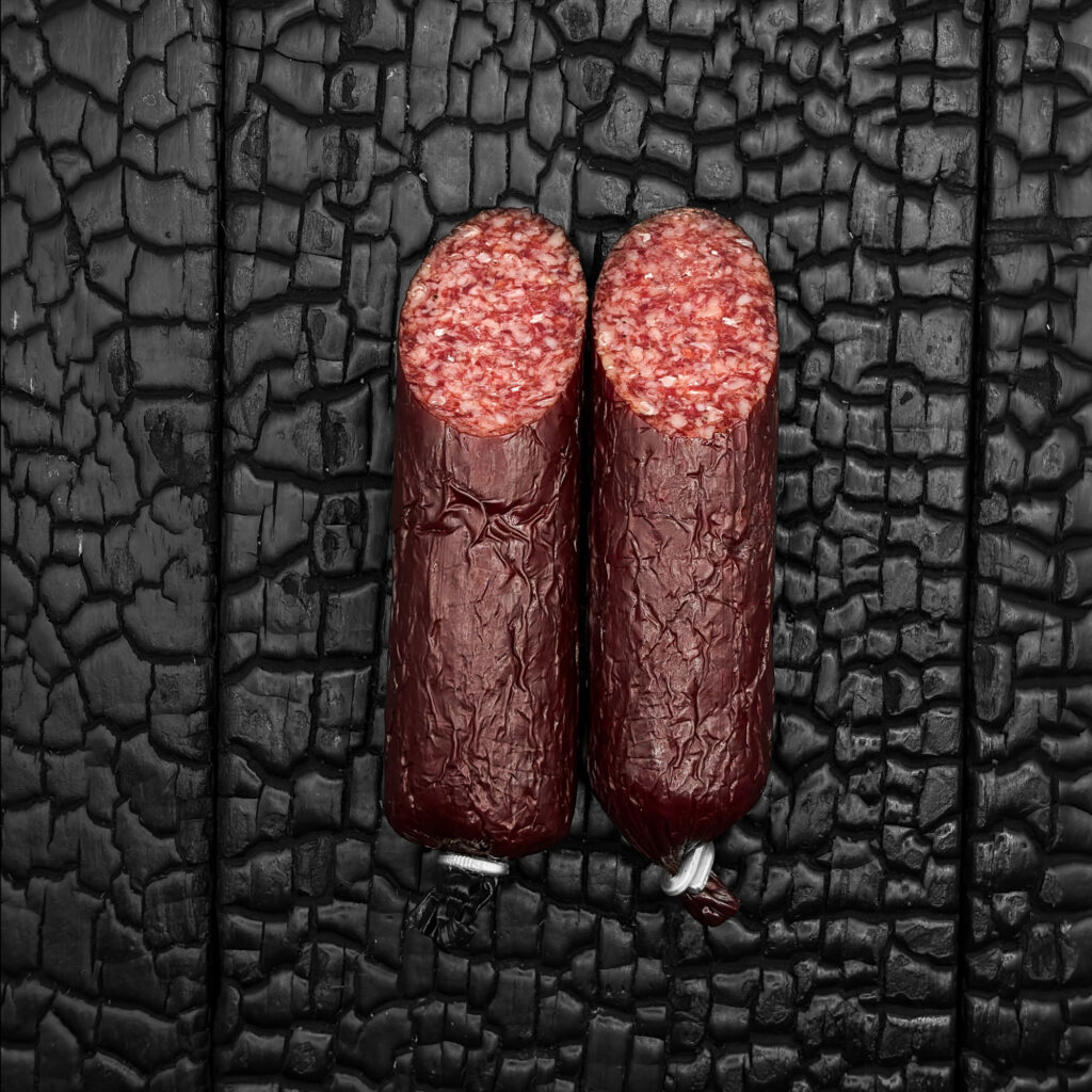 Zwei Wagyu-Salami-Stangen mit dunkelroter, leicht runzliger und glänzender Hülle, parallel nebeneinander platziert. Beide sind oben schräg angeschnitten und zeigen ein fein gekörntes Inneres mit gleichmäßig verteilten hellen Fettanteilen. Die Salami liegt mittig auf einer dunklen, verkohlten Holzoberfläche.