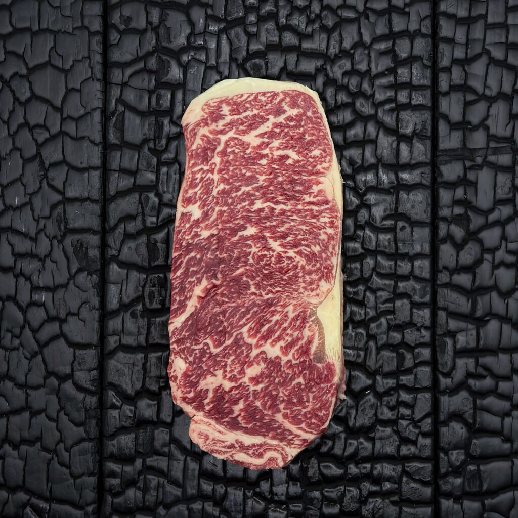 Rohes Strip Loin Steak vom Wagyu-Rind, stark marmoriert, rote Fleischfarbe mit hellem Fettrand, mittig auf dunkler, verkohlter Holzoberfläche.