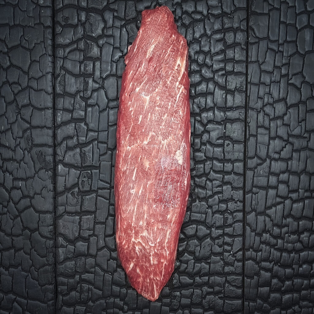 Rohes Teres-Major-Steak vom Wagyu-Rind, längliche Form, gut marmoriert, rote Farbe, mittig auf dunkler, verkohlter Holzoberfläche.
