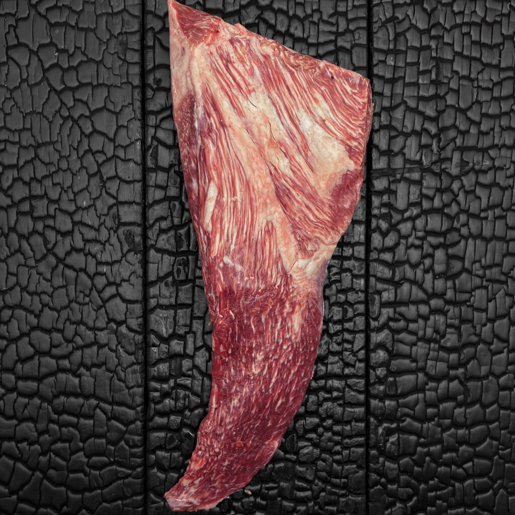 Rohes Tri-Tip im Ganzen vom Wagyu-Rind, gut marmoriert, rote Fleischfarbe mit grober Faserstruktur und Fettdeckel, länglich-dreieckig geformt, mittig auf dunkler, verkohlter Holzoberfläche.