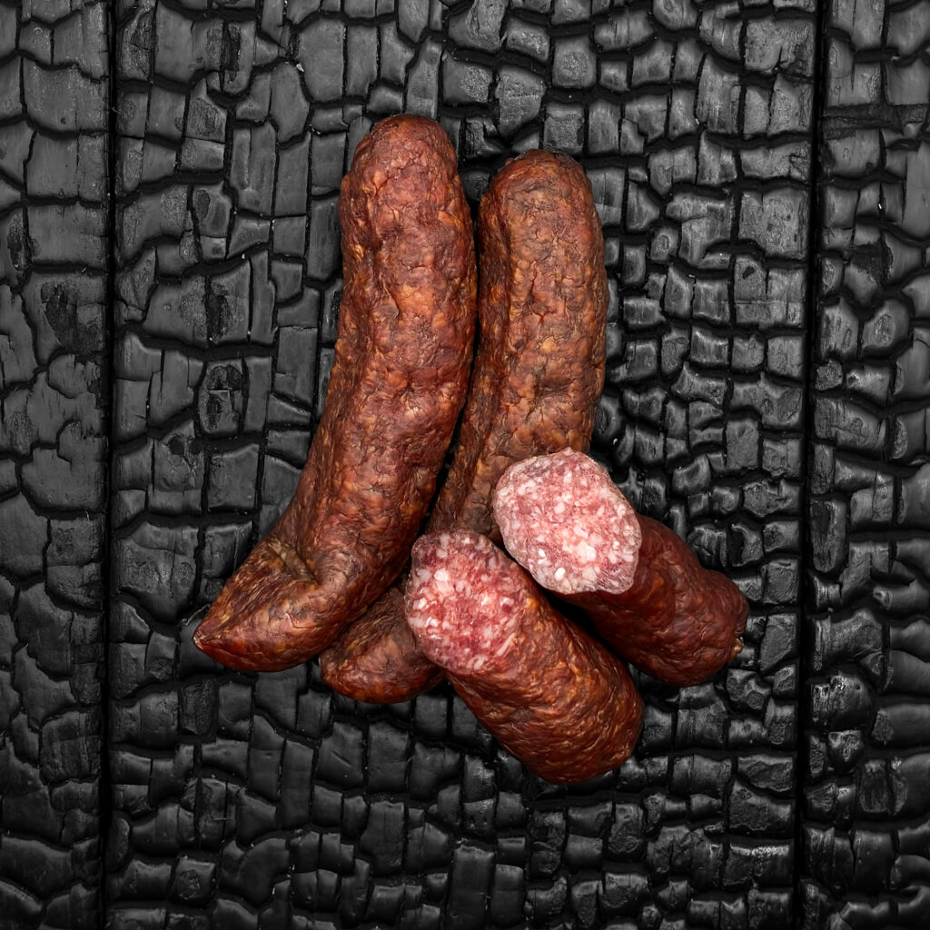Mehrere Wagyu Jausenwürstel mit Trüffel und Steinpilz, mit dunkelbrauner, leicht glänzender Wursthülle, locker übereinander angeordnet. Zwei Stück sind quer angeschnitten und zeigen ein fein gekörntes, rötliches Inneres mit hellen Fettanteilen. Die Würstel liegen mittig auf einer dunklen, verkohlten Holzoberfläche.
