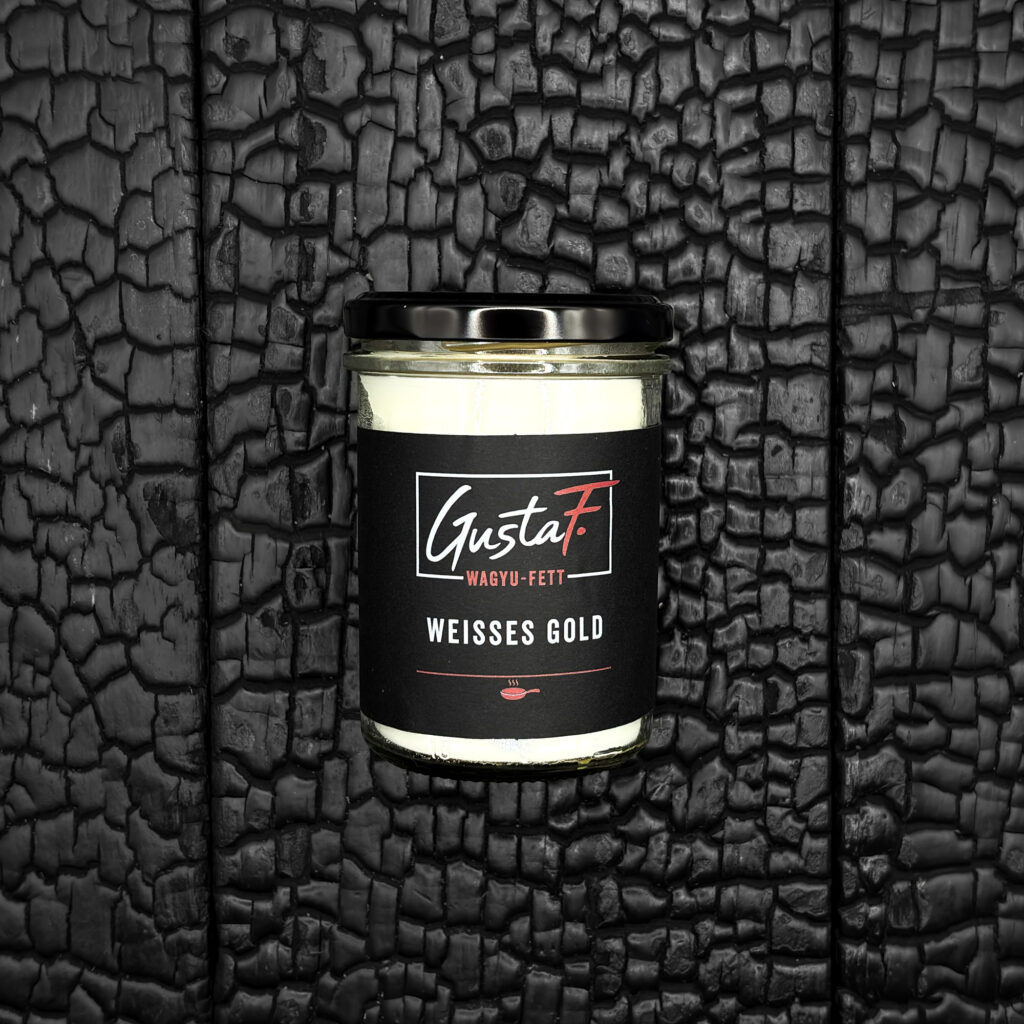 Ein Glas mit Wagyu-Rinderfett, gefüllt mit einer hellen, cremeweißen Masse. Das Glas hat einen schwarzen Schraubdeckel und ein dunkles Etikett mit der Aufschrift „GustaF. Wagyu-Fett – Weißes Gold“. Es liegt mittig auf einer dunklen, verkohlten Holzoberfläche.